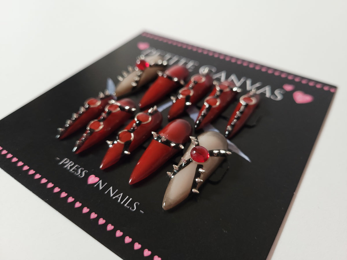 Close-up of REBIRTH long stiletto press-on nails — red and nude with metal spikes, chains, rings and red gems
